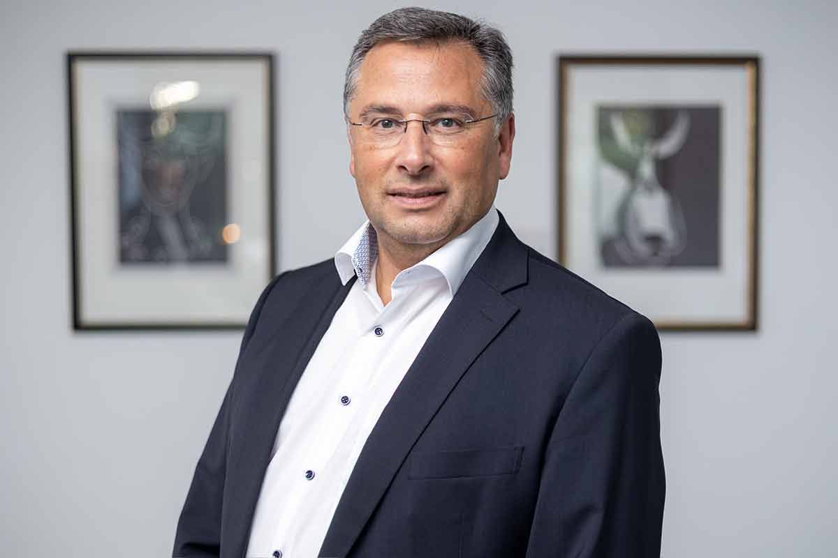 Ein Mann in Anzug und weißem Hemd steht vor zwei gerahmten Bildern.