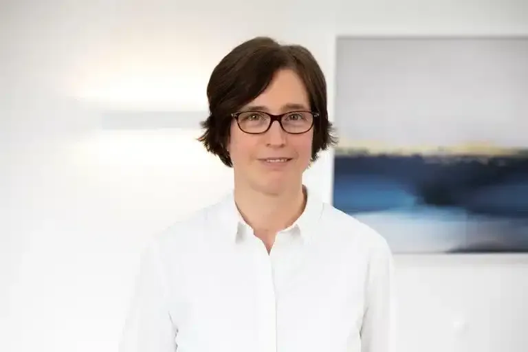 Eine Person mit kurzen dunklen Haaren und Brille in einem weißen Hemd steht vor einem verschwommenen Wandgemälde.