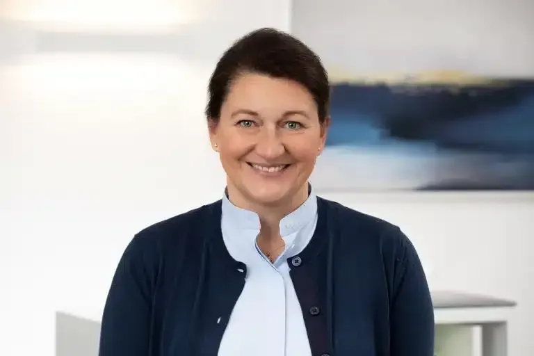 Lächelnde Frau in marineblauer Jacke und weißer Bluse steht in einem Innenraum vor verschwommenem Hintergrund