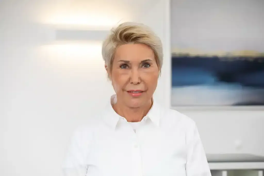 Porträt einer Person in einem weißen Hemd, die in einem hellen Büro steht, im Hintergrund ist ein Wandgemälde zu sehen.
