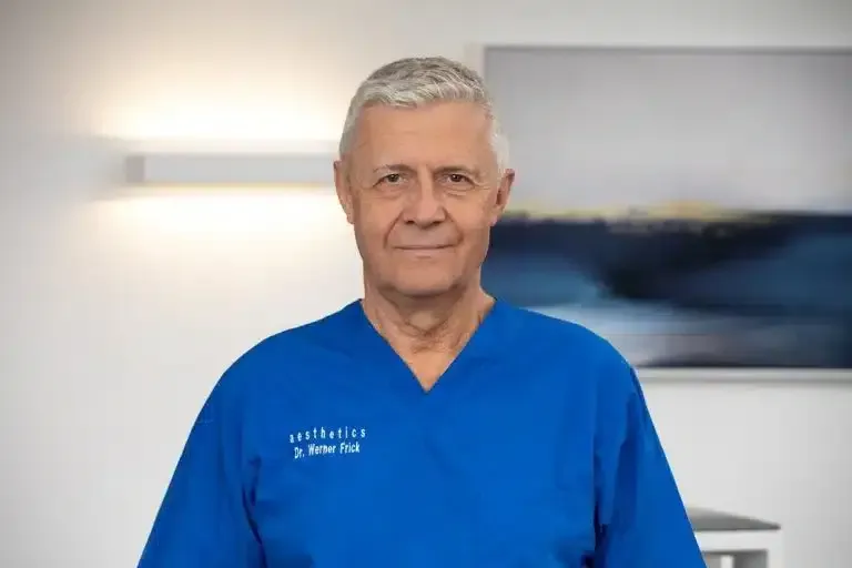 Ein Mann in blauer OP-Kleidung steht in einem Innenraum vor einem verschwommenen Wandgemälde und blickt in die Kamera.