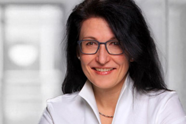 Lächelnde Frau mit schwarzen Haaren und Brille, die eine weiße Bluse trägt, vor einem leicht verschwommenen Hintergrund