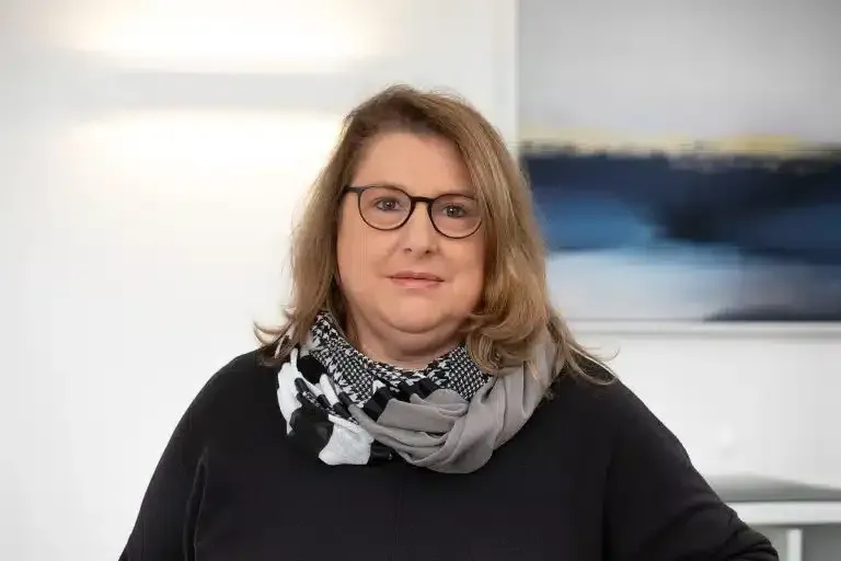 Porträt einer Frau mit Brille und gemustertem Schal, die in einem Innenraum vor einem verschwommenen Hintergrund steht