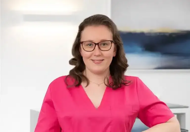 Frau in leuchtend pinkfarbener Bluse und Brille, lächelnd vor einem verschwommenen, büroähnlichen Hintergrund.