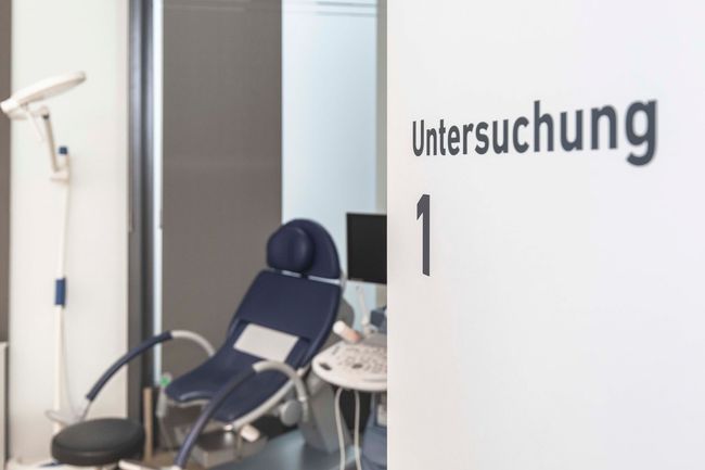 Leerer Untersuchungsraum mit einem blauen Liegestuhl und dem Schild „Untersuchung 1“