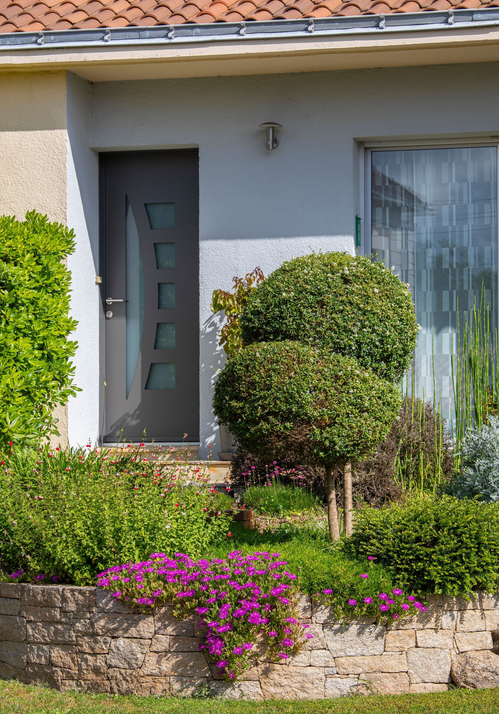 Cour avant avec arbustes bien entretenus, porte et mur de soutènement en pierre avec des fleurs violettes.