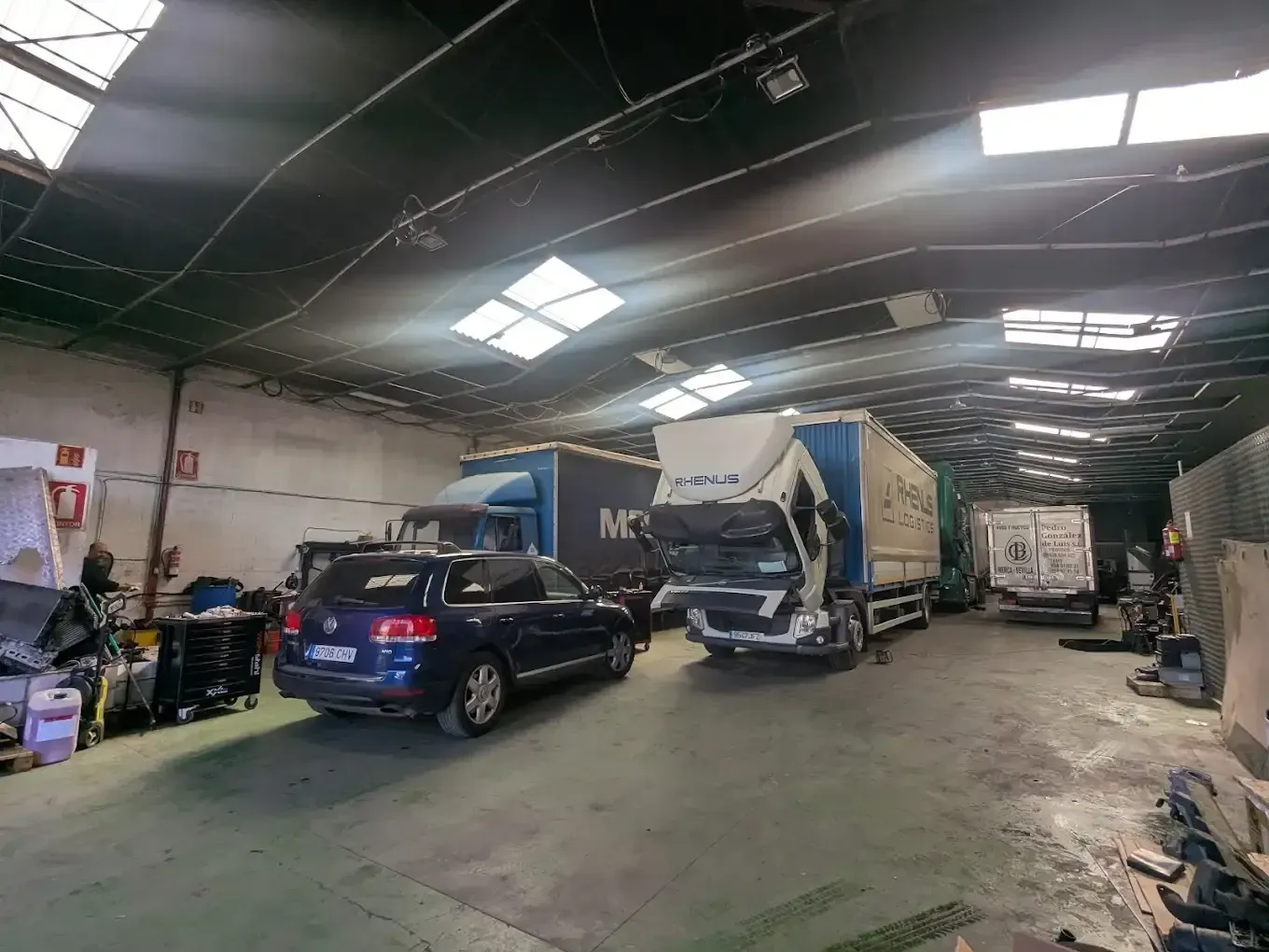 Dentro de un almacén: un coche y camiones estacionados. Se filtra algo de luz natural por las aberturas del techo.