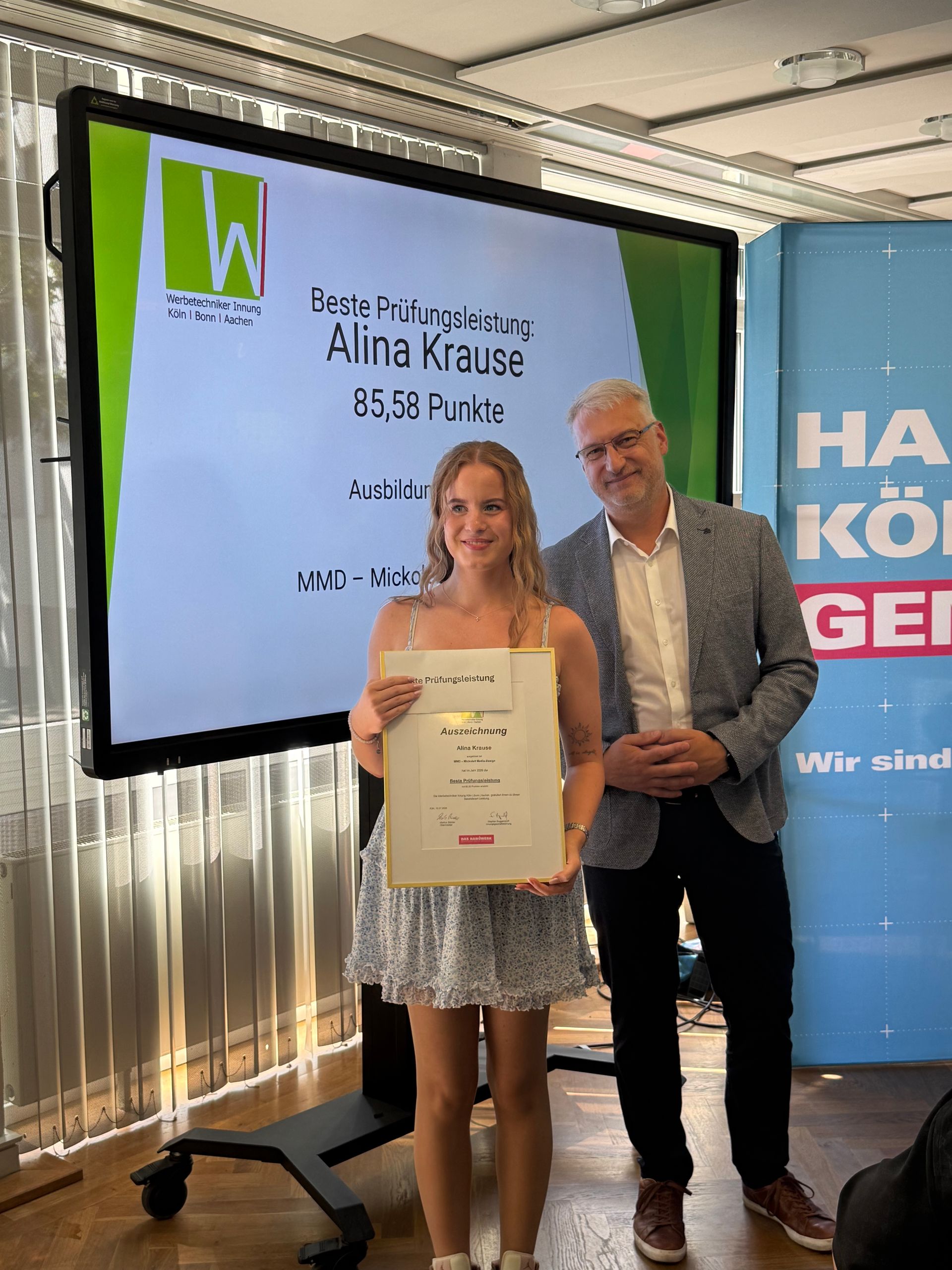 Zwei Personen halten auf einer Bühne neben einer großen Präsentationsleinwand eine Urkunde in die Höhe.