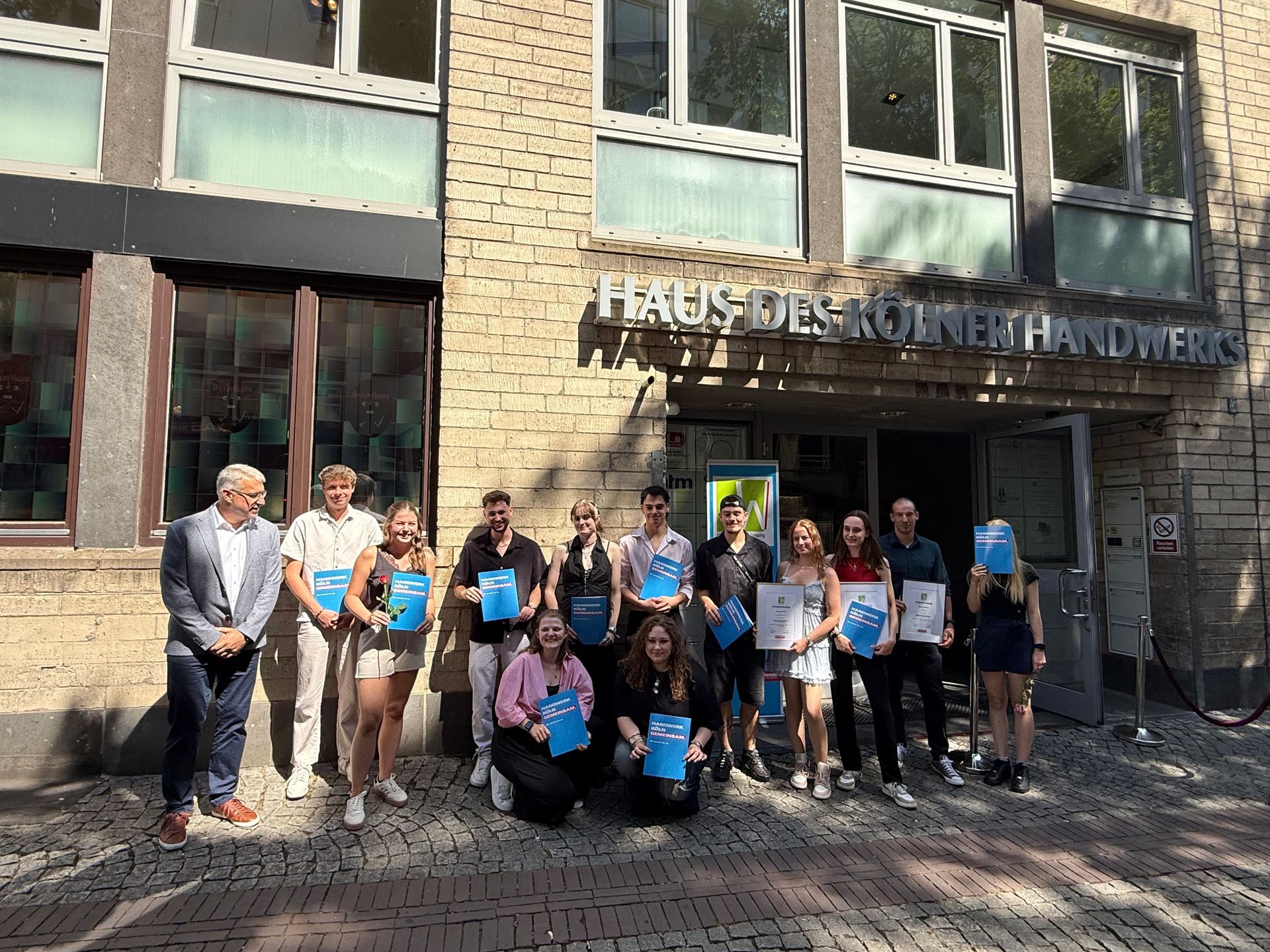 Eine Gruppe von Menschen posiert vor dem Eingang eines Baumarkts auf einer sonnigen Straße.