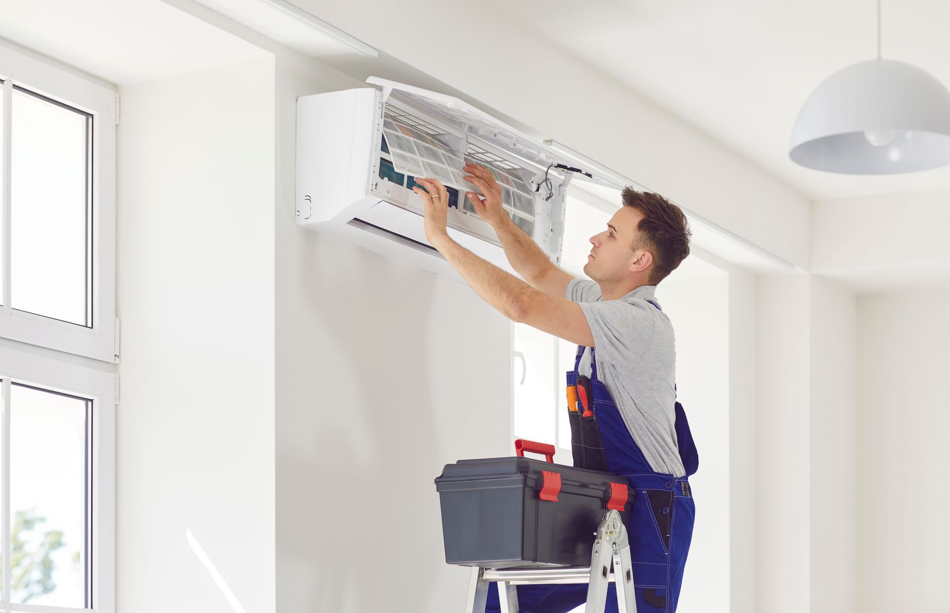 Un homme en salopette bleue nettoie un filtre de climatiseur, debout sur une échelle avec une boîte à outils dans une pièce blanche.