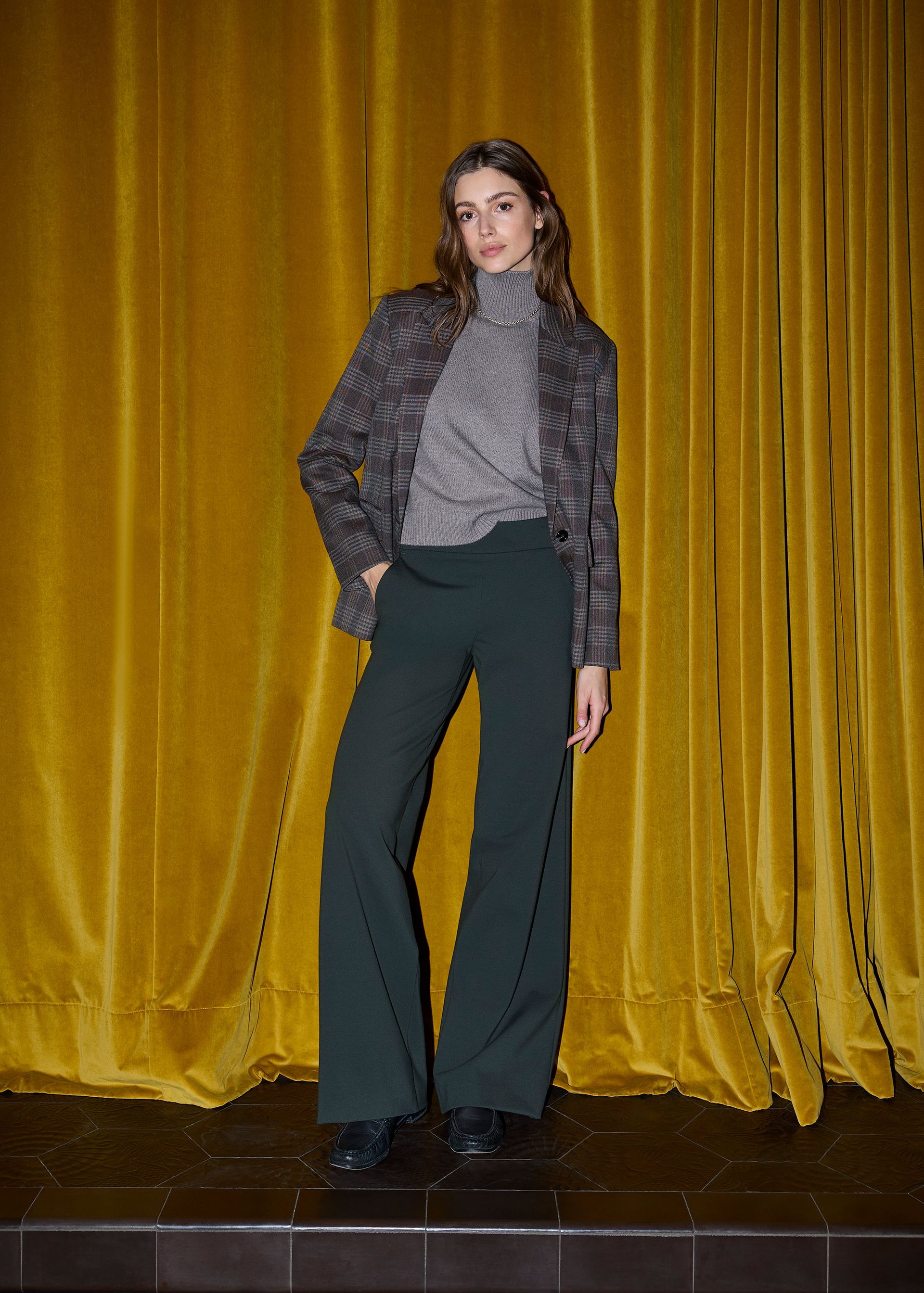 Frau in grauem Rollkragenpullover, geblümtem Blazer und grüner Hose posiert vor einem goldenen Vorhang.