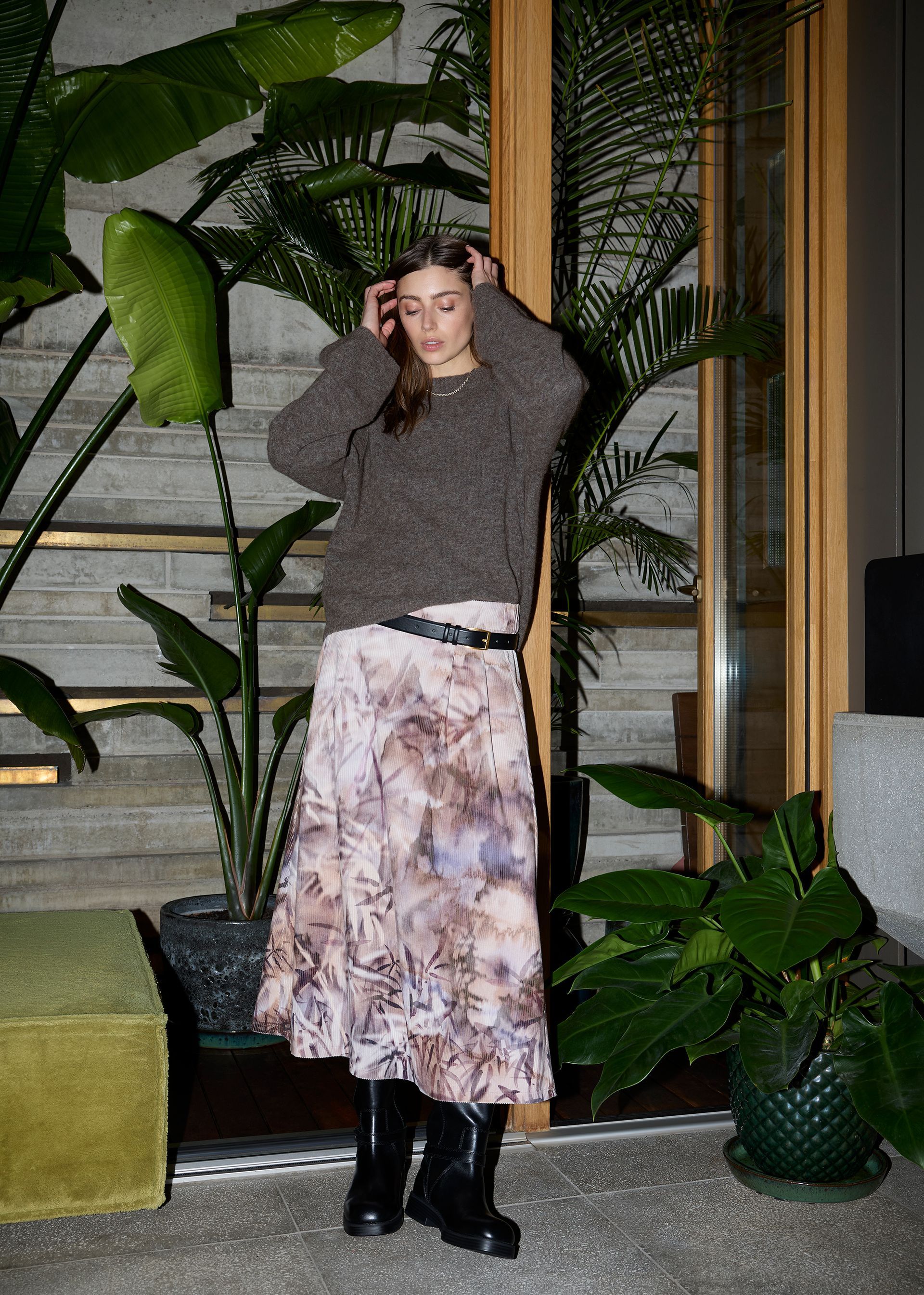 Frau in Pullover und geblümtem Rock, berührt ihr Haar, in der Nähe von Pflanzen und einer Holztür.