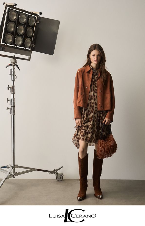 Frau im Herbstoutfit, posiert vor einer Studioleuchte. Sie trägt eine Jacke, ein gemustertes Kleid, Stiefel und hält eine Tasche.