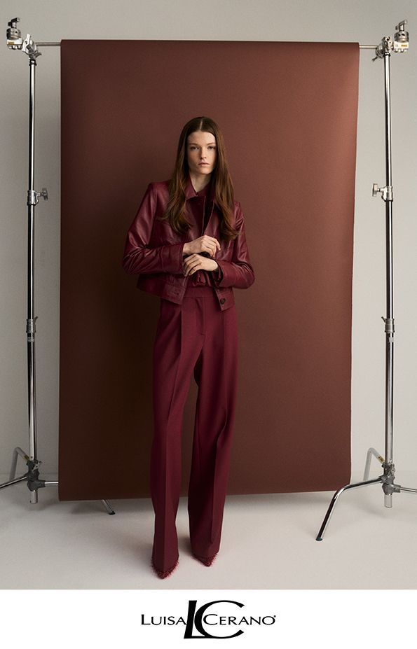 Frau in burgunderfarbenem Hosenanzug und Lederjacke posiert vor einem braunen Hintergrund.