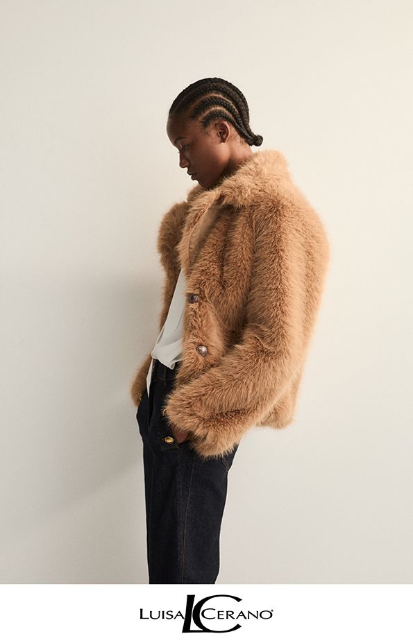 Eine Person in einer braunen, zotteligen Jacke, einem weißen Hemd und einer dunklen Hose steht vor einer hellen Wand.