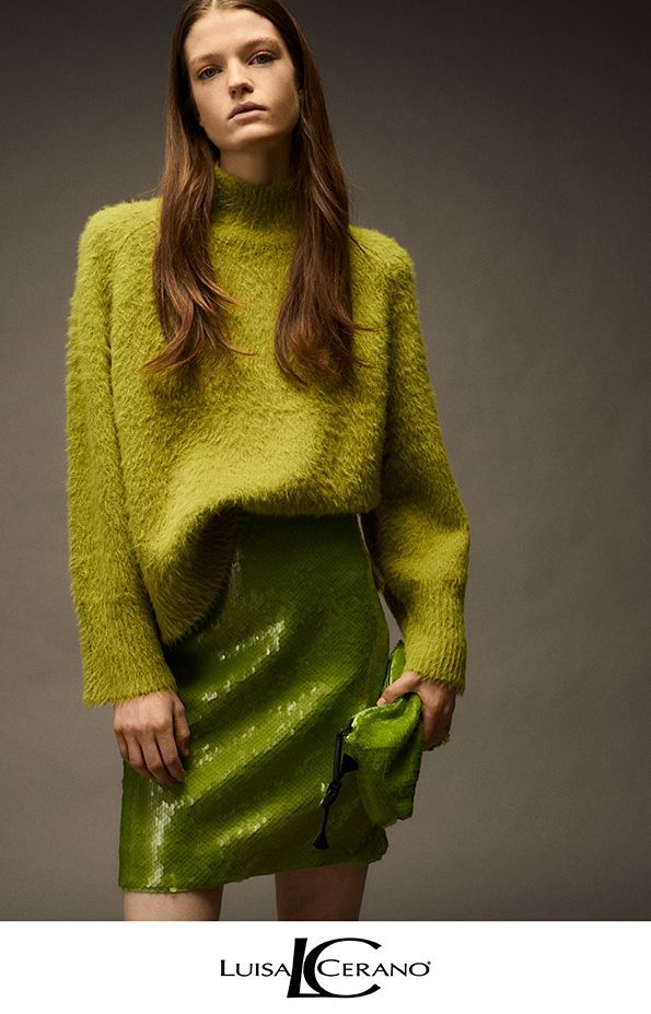 Frau in hellgrünem Pullover und Paillettenrock, die eine Tasche hält, vor grauem Hintergrund.