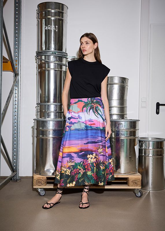 Frau in schwarzem Oberteil und buntem Rock posiert neben Metallfässern.