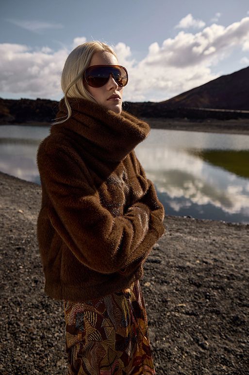 Eine Person mit Sonnenbrille und braunem Pullover steht an einem Seeufer; im Hintergrund sind Berge und Wolken zu sehen.