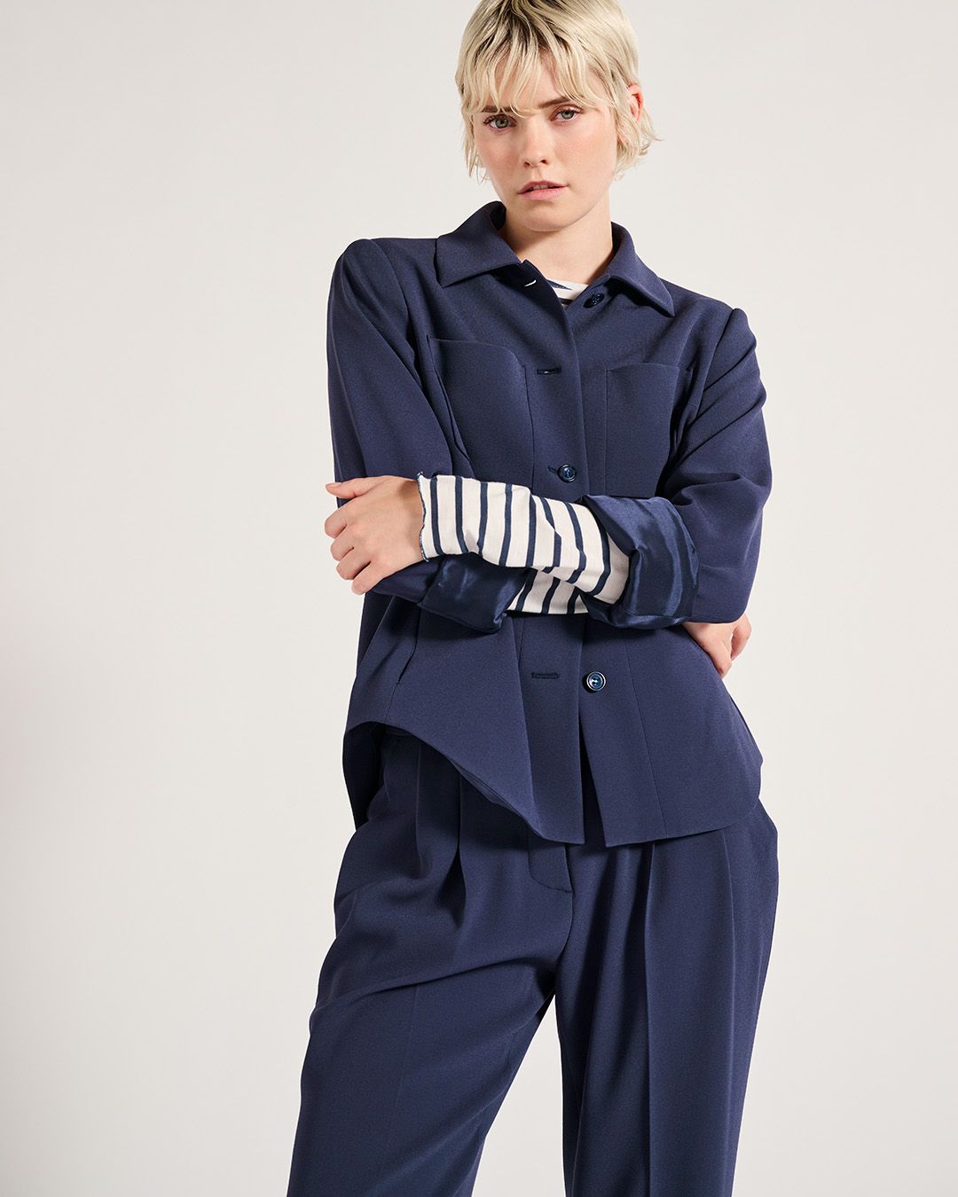 Frau in einem marineblauen Anzug mit einem gestreiften Hemd darunter, die Arme verschränkt.
