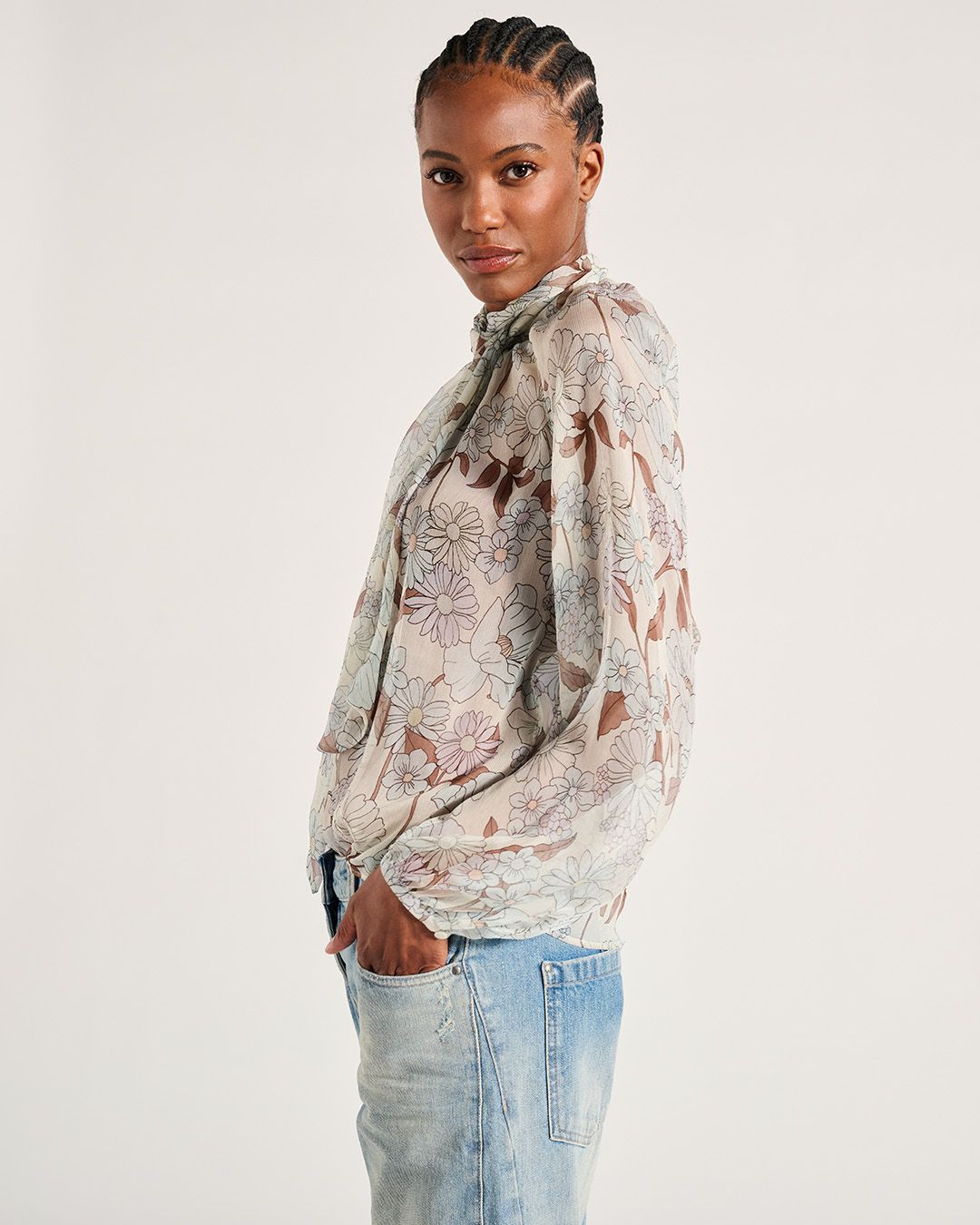 Frau trägt eine Bluse mit Blumenmuster und Jeans, die Hände in den Taschen, und schaut zur Seite.