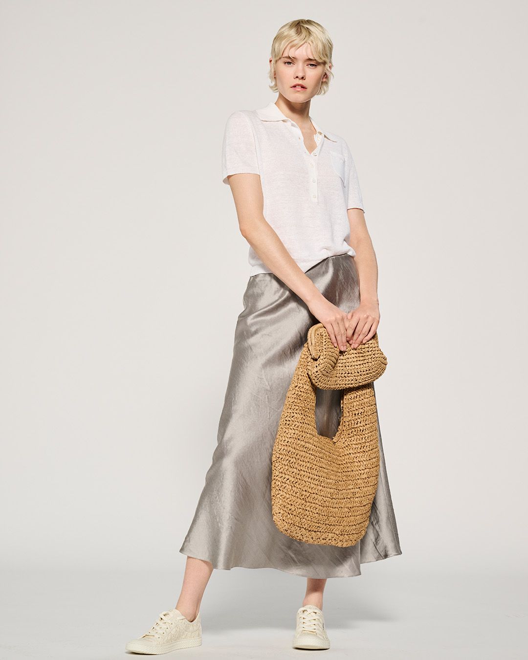 Frau in weißem Top, silbernem Rock und Turnschuhen hält eine gewebte Tasche und posiert in einem Studio.