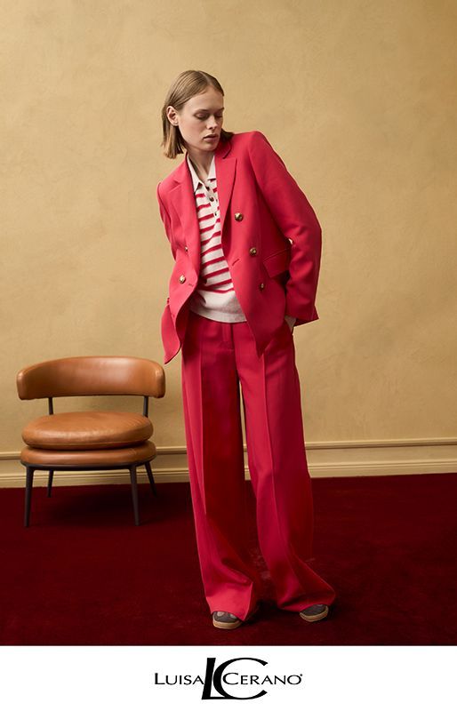 Frau in rotem Blazer und Hose, gestreiftem Hemd, steht auf einem roten Teppich neben einem Stuhl und einer beigen Wand.