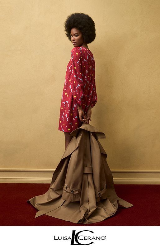 Frau in rotem Blumenkleid, die einen beigen Mantel hält, steht auf einem roten Teppich vor einer gelben Wand.