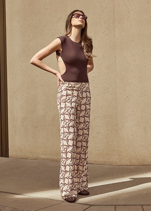 Frau in braunem Oberteil und gemusterter Hose, im Freien, Sonnenbrille, Hände in die Hüften gestemmt.