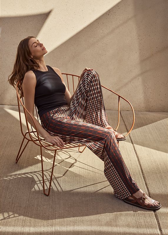 Frau in gemusterter Hose und schwarzem Oberteil sitzt auf einem Metallstuhl und genießt die Sonne.
