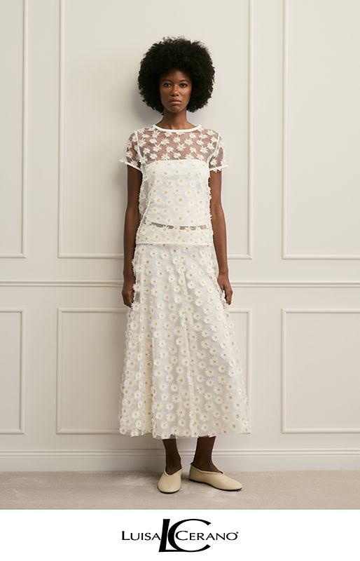 Ein Model trägt einen weißen, geblümten, zweiteiligen Midirock und ein passendes Oberteil vor einer weißen, getäfelten Wand.