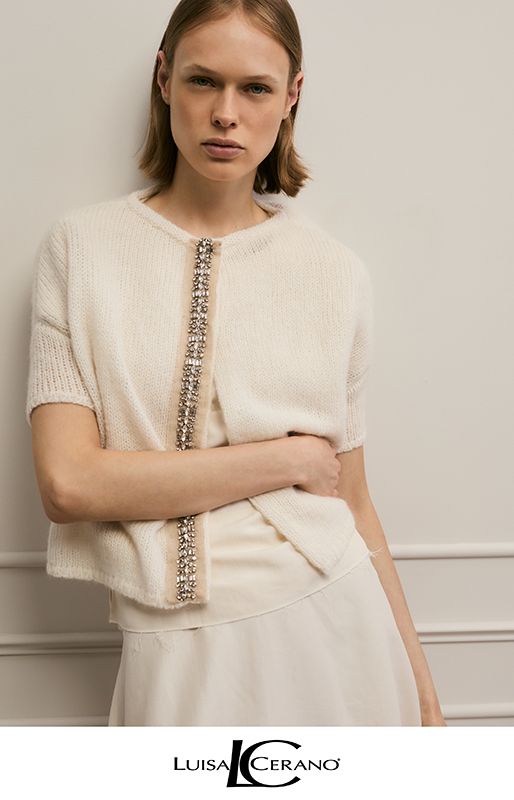 Eine Person, die eine elfenbeinfarbene, kurzärmelige Strickjacke mit juwelenbesetzter Knopfleiste über einem passenden Oberteil und Rock trägt.