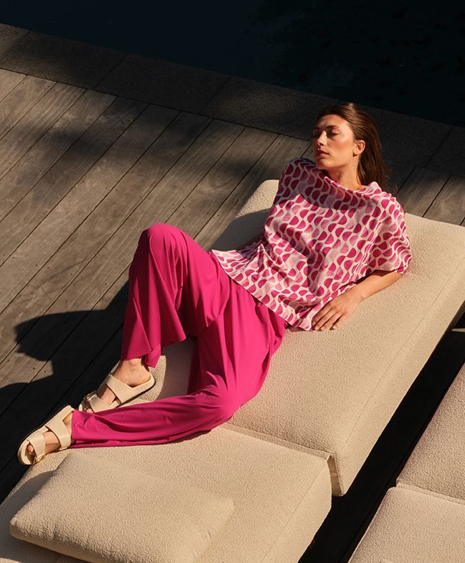 Eine Person in einem rosa gemusterten Oberteil und einer pinkfarbenen Hose liegt in der Sonne auf einer beigen Sonnenliege auf einer Holzterrasse.