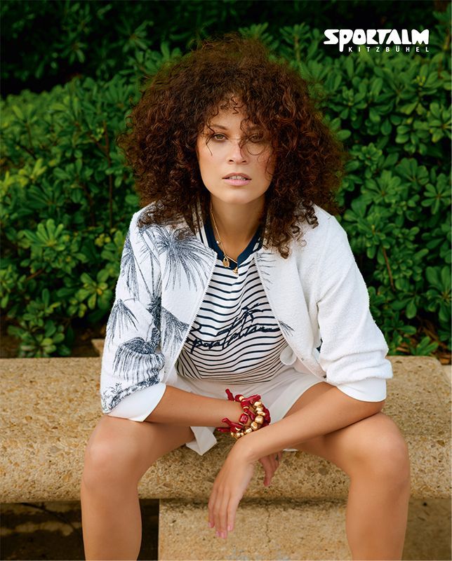 Eine Person mit lockigem Haar sitzt im Freien auf einer Steinbank. Sie trägt eine weiße gemusterte Jacke und ein gestreiftes Hemd.