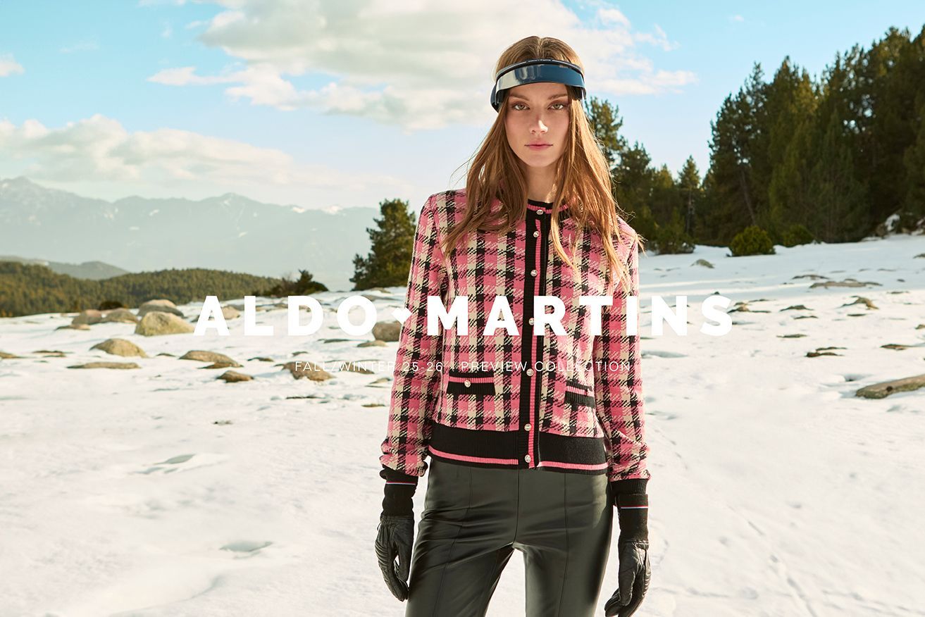 Frau in rosa Tweedjacke und Skibrille auf einem schneebedeckten Berg.