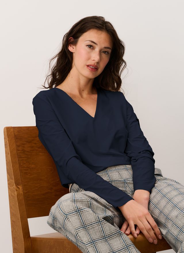 Frau in marineblauem Oberteil und karierter Hose sitzt auf einem Holzstuhl und blickt in die Kamera.