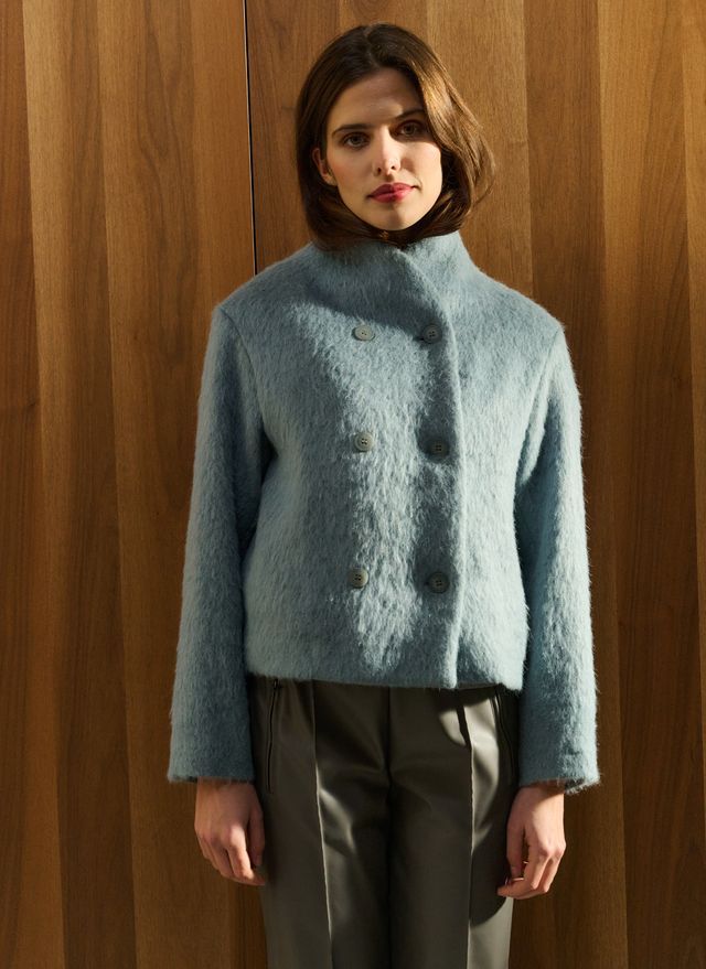 Frau in hellblauer Jacke und grauer Hose steht vor einer Holzwand.