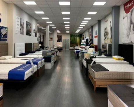 Pasillo de una tienda de muebles con filas de camas y colchones bajo luces brillantes en el techo