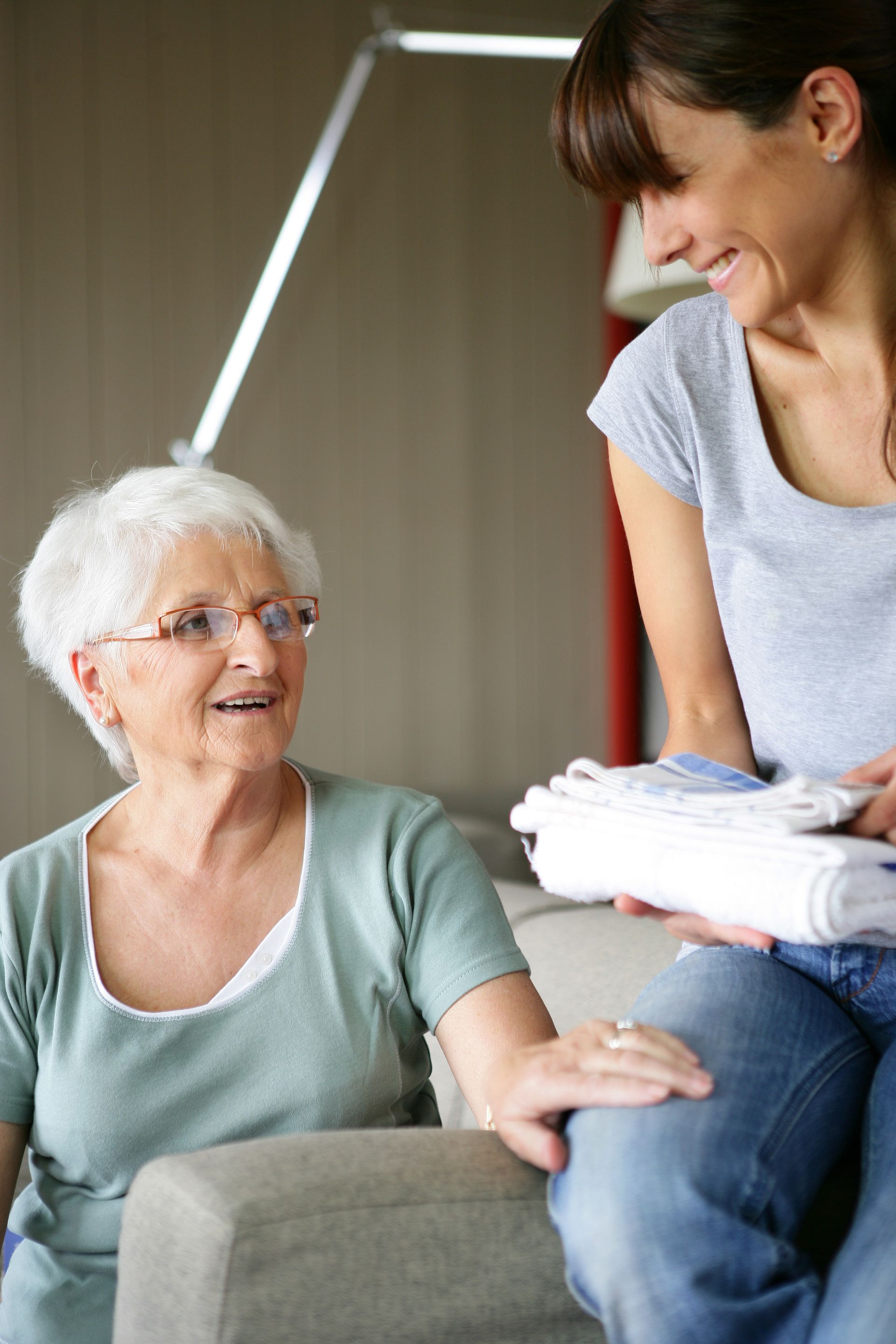 Une femme sourit, plie des vêtements et les offre à une femme âgée souriante, assise sur un canapé. À l'intérieur.