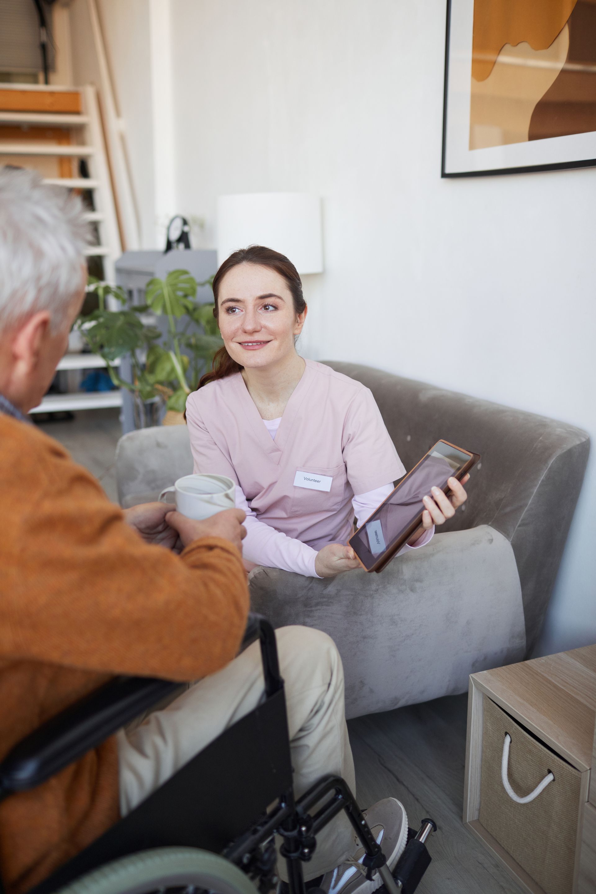 Un soignant avec une tablette parle à un homme âgé en fauteuil roulant, souriant dans un salon.