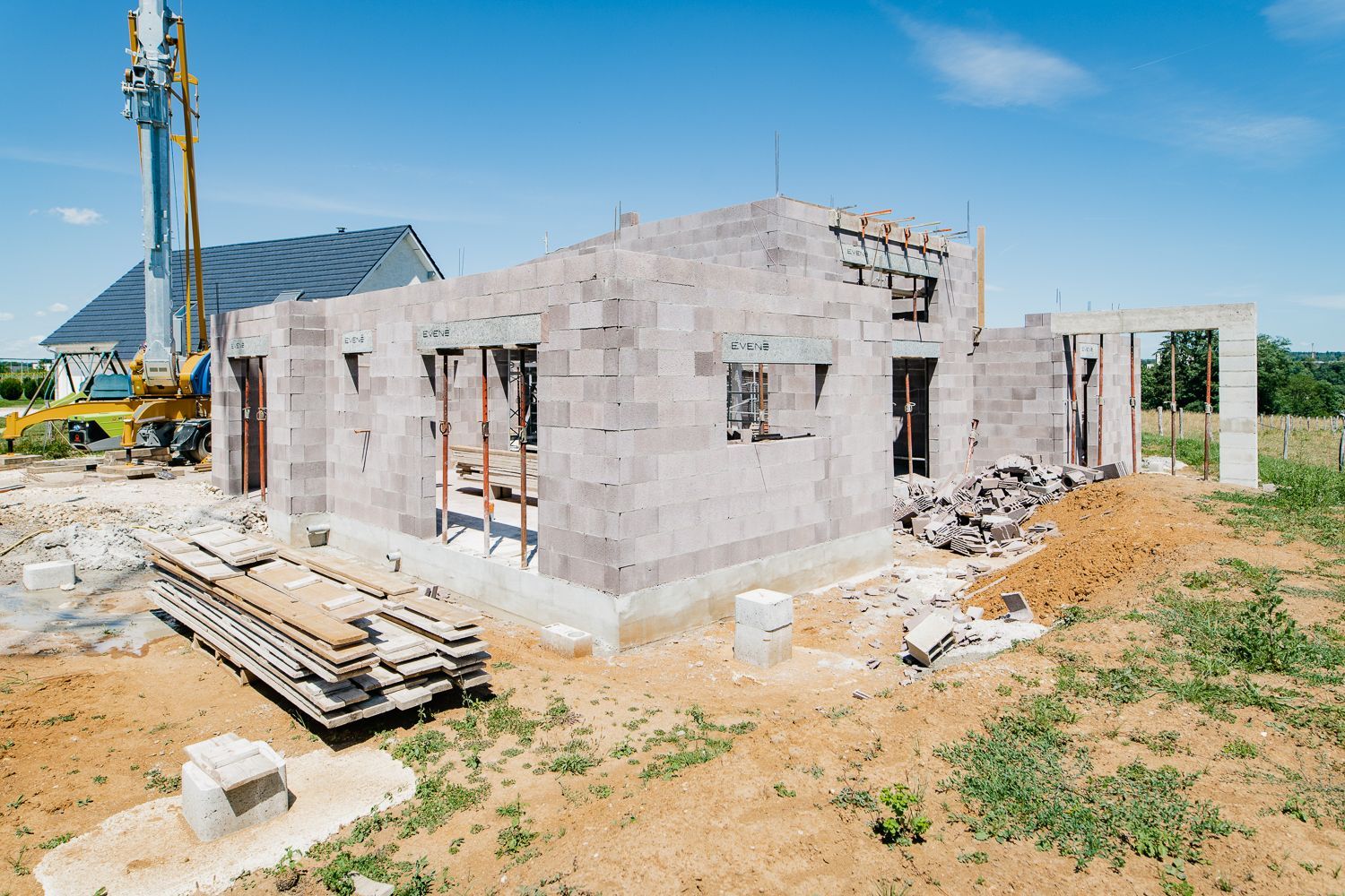 Chantier : une ossature de maison en blocs gris, ciel bleu, grue jaune en arrière-plan.