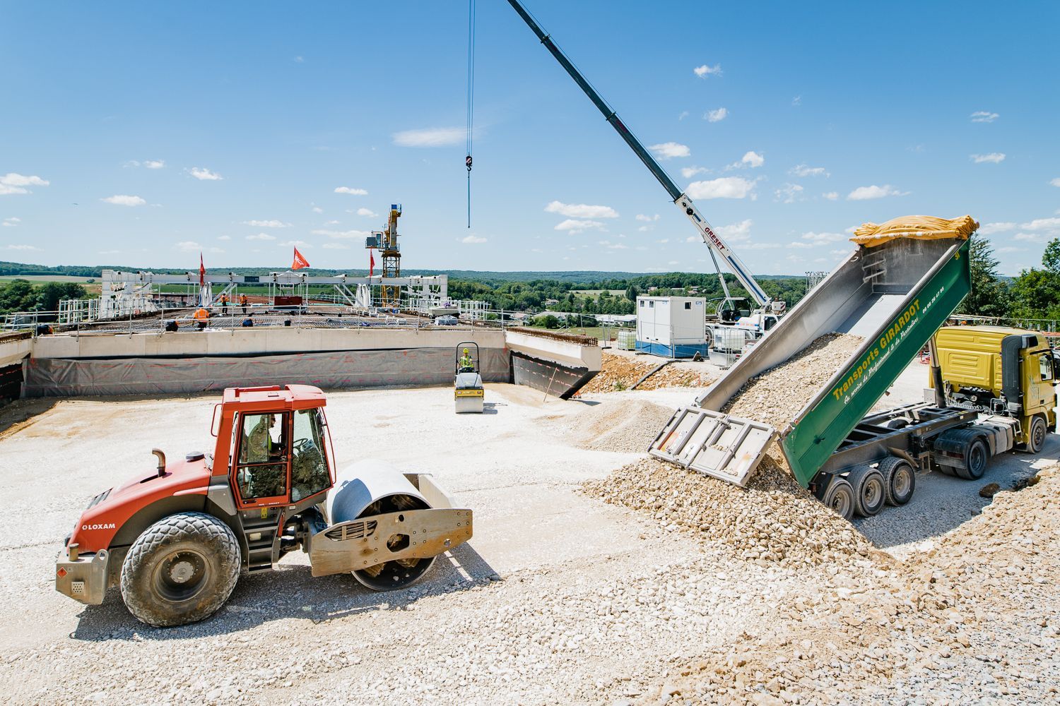 Camion-benne déchargeant du gravier, rouleau compactant le sol, grue en arrière-plan