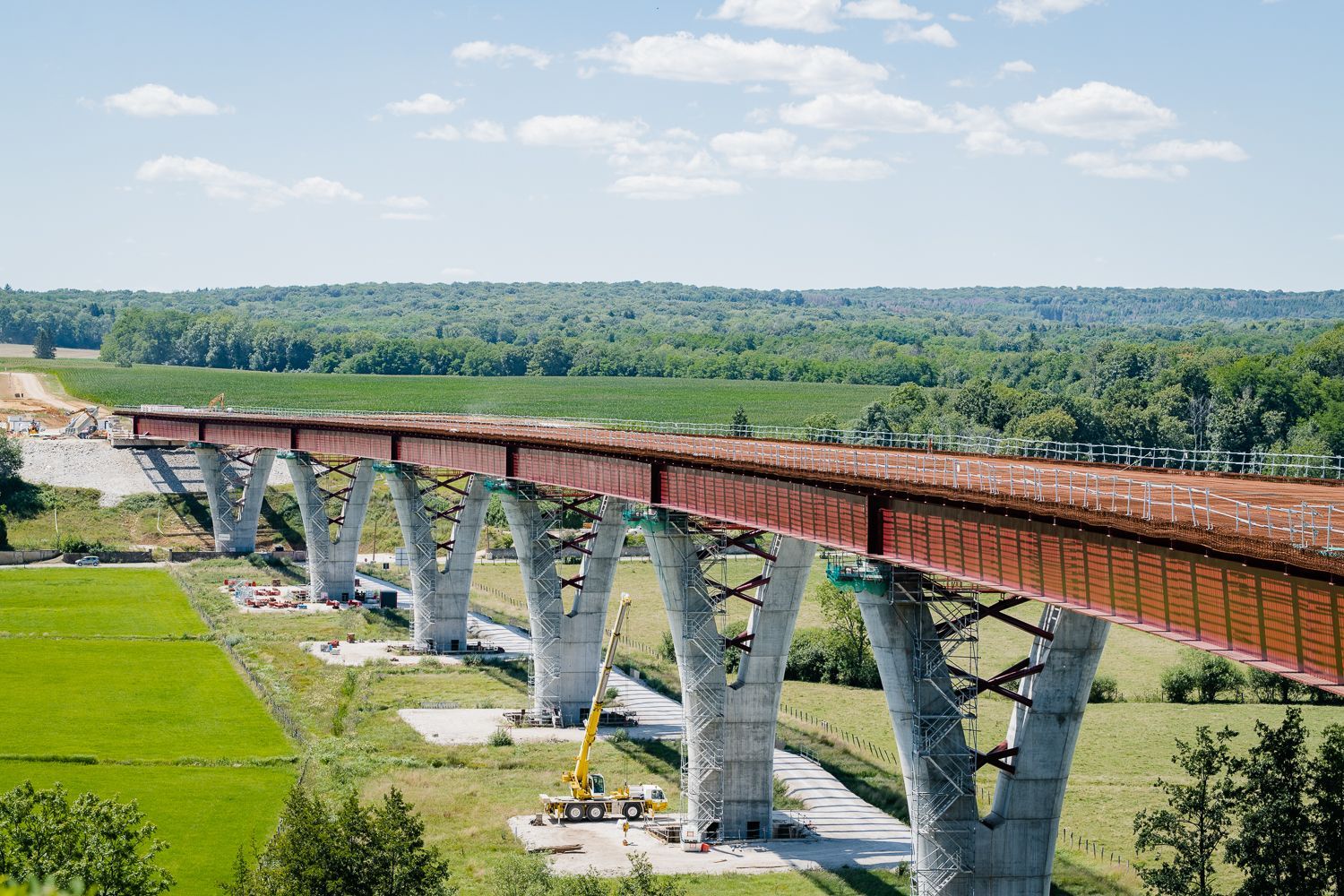 Pont en construction enjambant une vallée, avec des piliers en béton et un tablier en acier brun-rouge