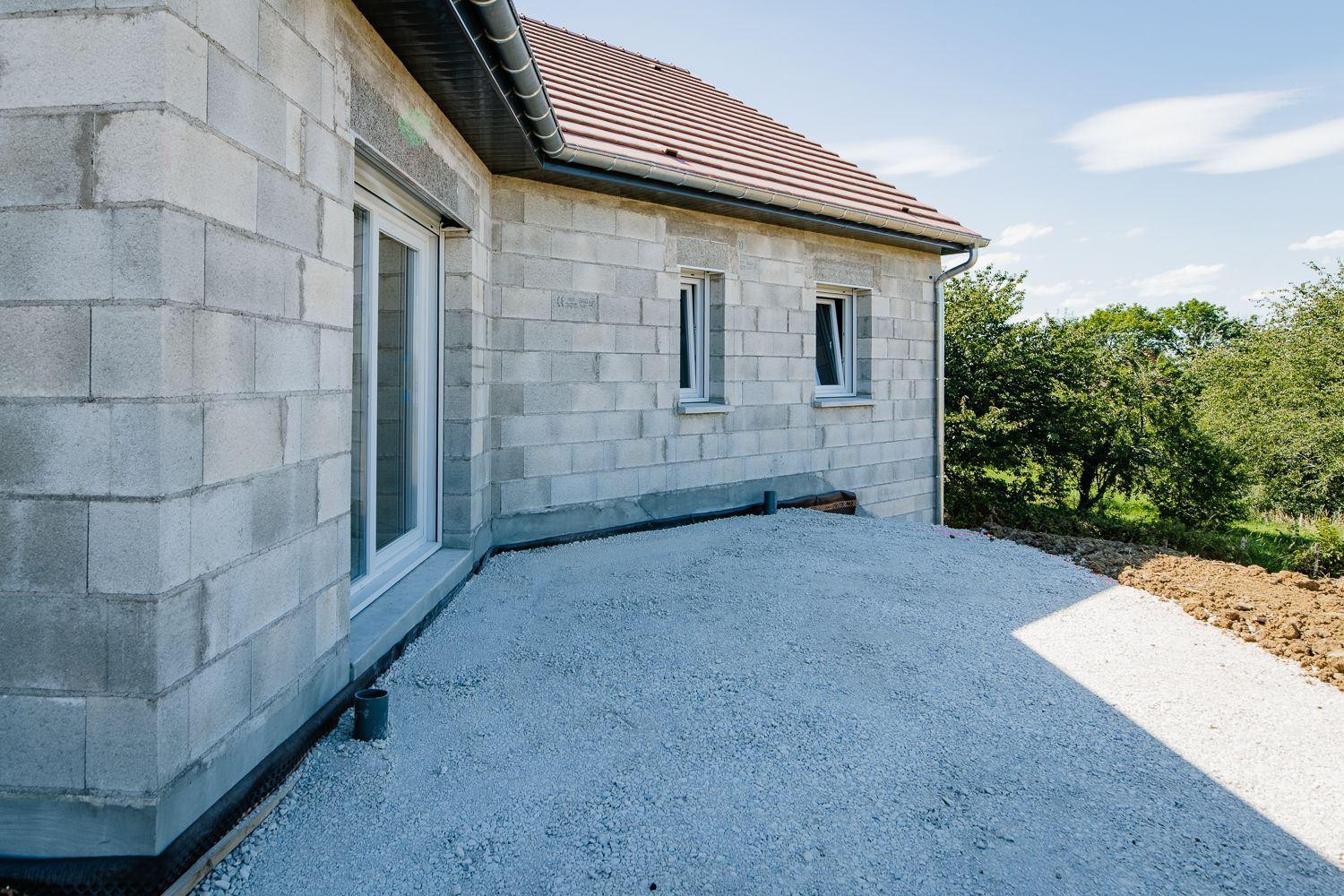 Maison en blocs de béton gris avec fenêtres à cadre blanc, patio en gravier et toit en tuiles rouges