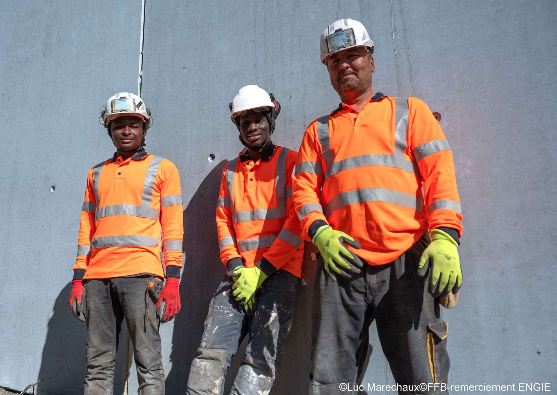 Trois ouvriers du bâtiment portant des chemises de sécurité orange et des casques debout devant un mur gris