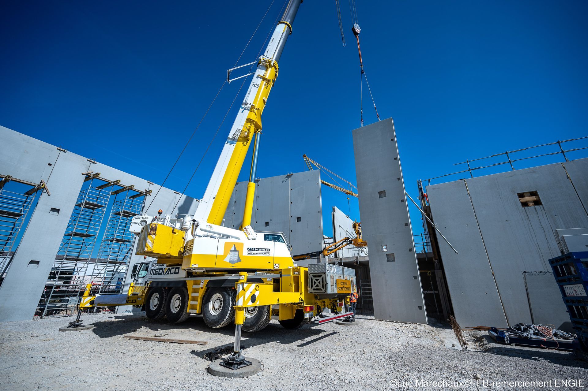 Une grue jaune soulevant un mur de béton en place sur un chantier de construction