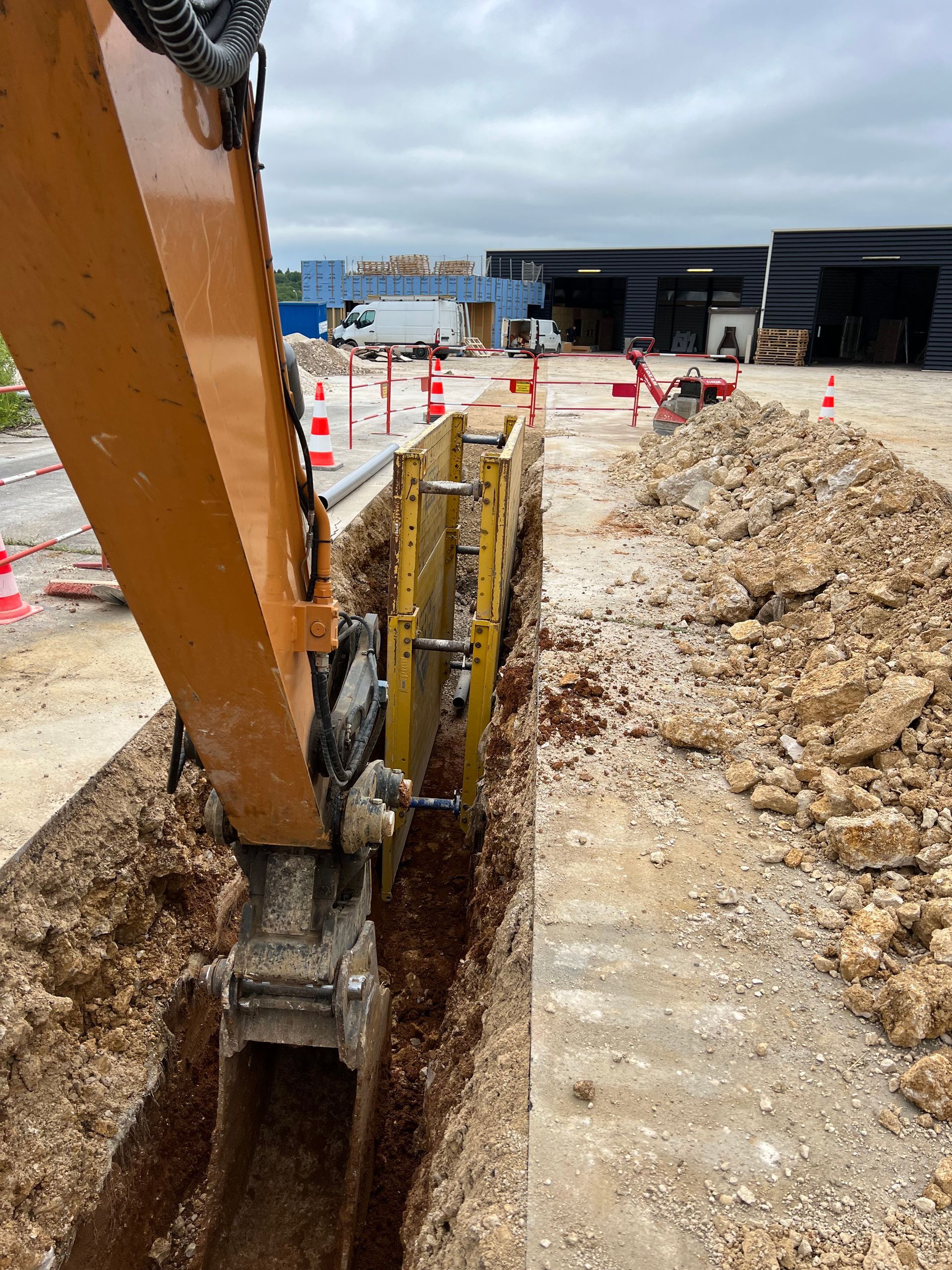Une excavatrice creuse une tranchée près d'un bâtiment, des cônes orange marquent la zone