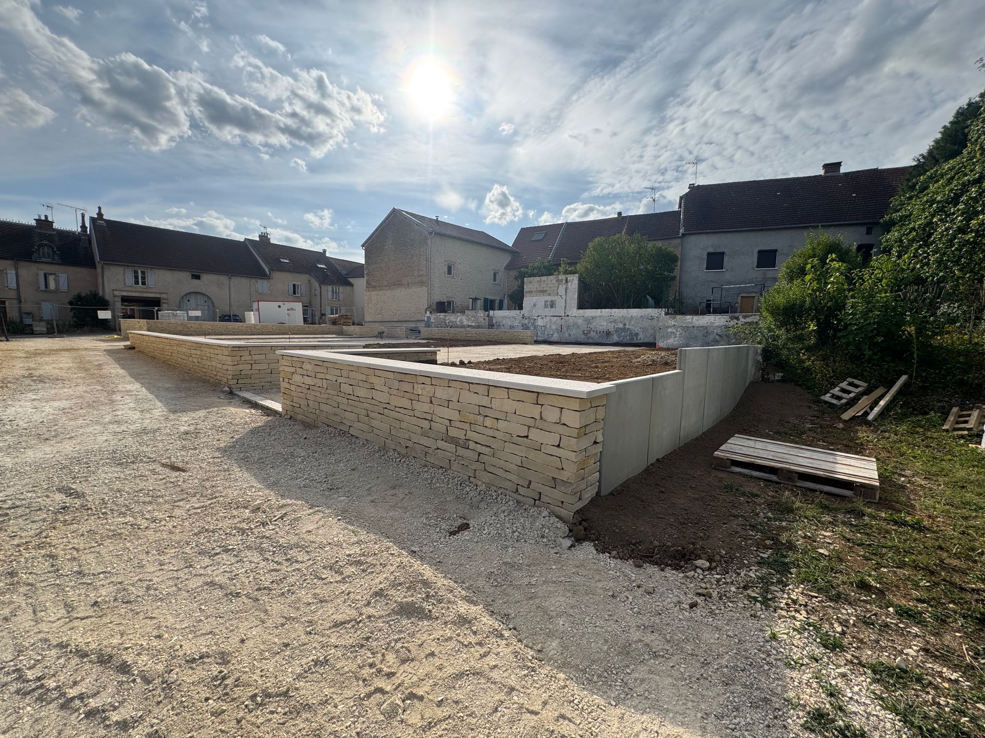 Chantier de construction avec murs en pierre, sol en gravier et bâtiments en arrière-plan sous un ciel ensoleillé