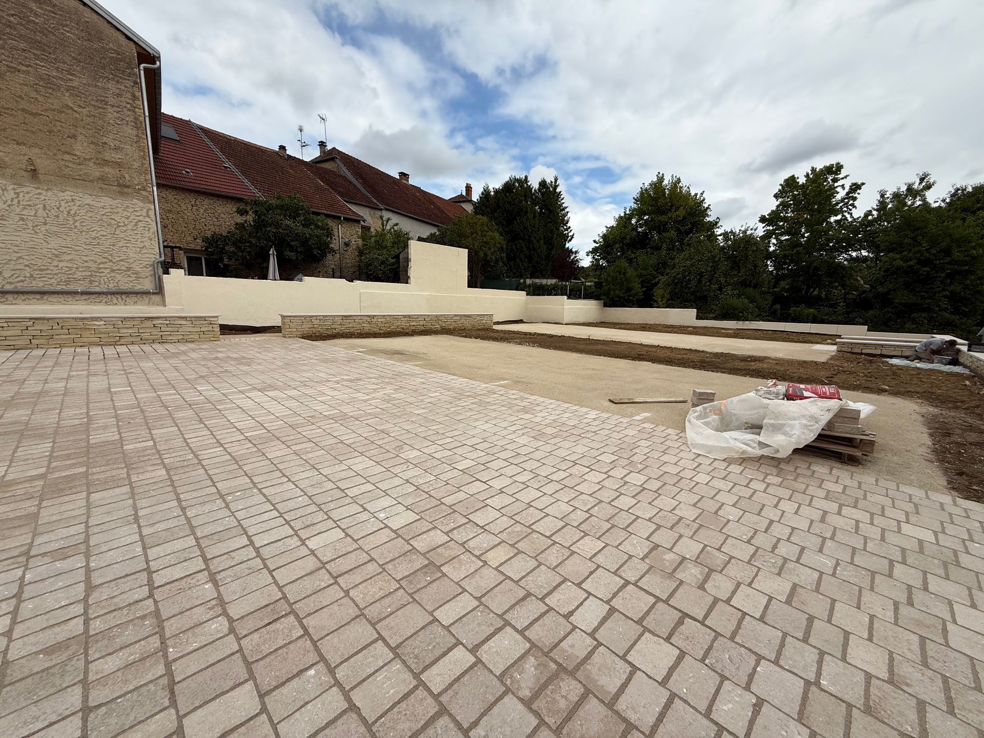 Espace extérieur nouvellement pavé avec des pavés beiges, des murets et des bâtiments sous un ciel nuageux