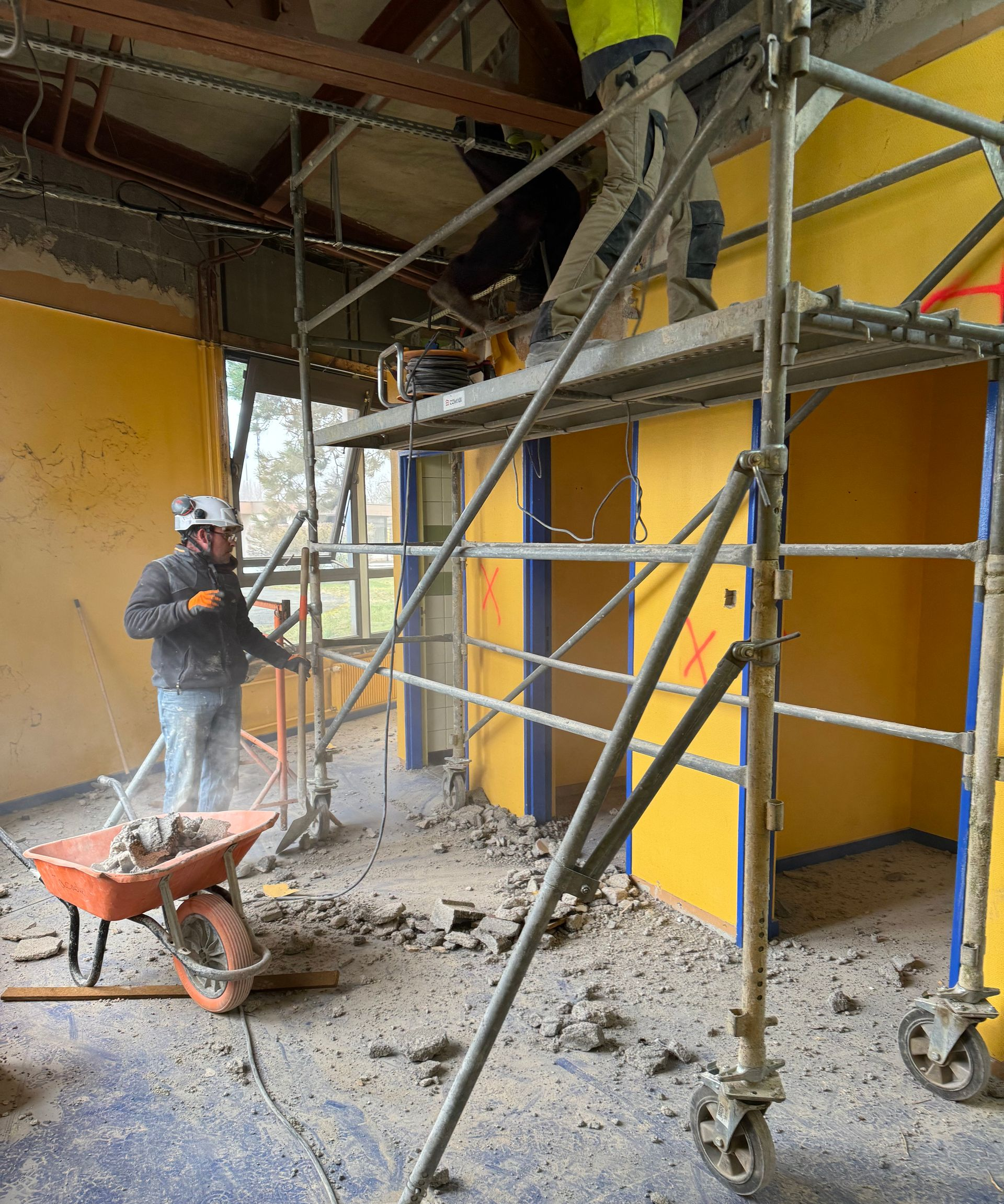 Des ouvriers du bâtiment démolissent une pièce ; l'un sur un échafaudage avec une scie, l'autre déblayant les débris avec une brouette