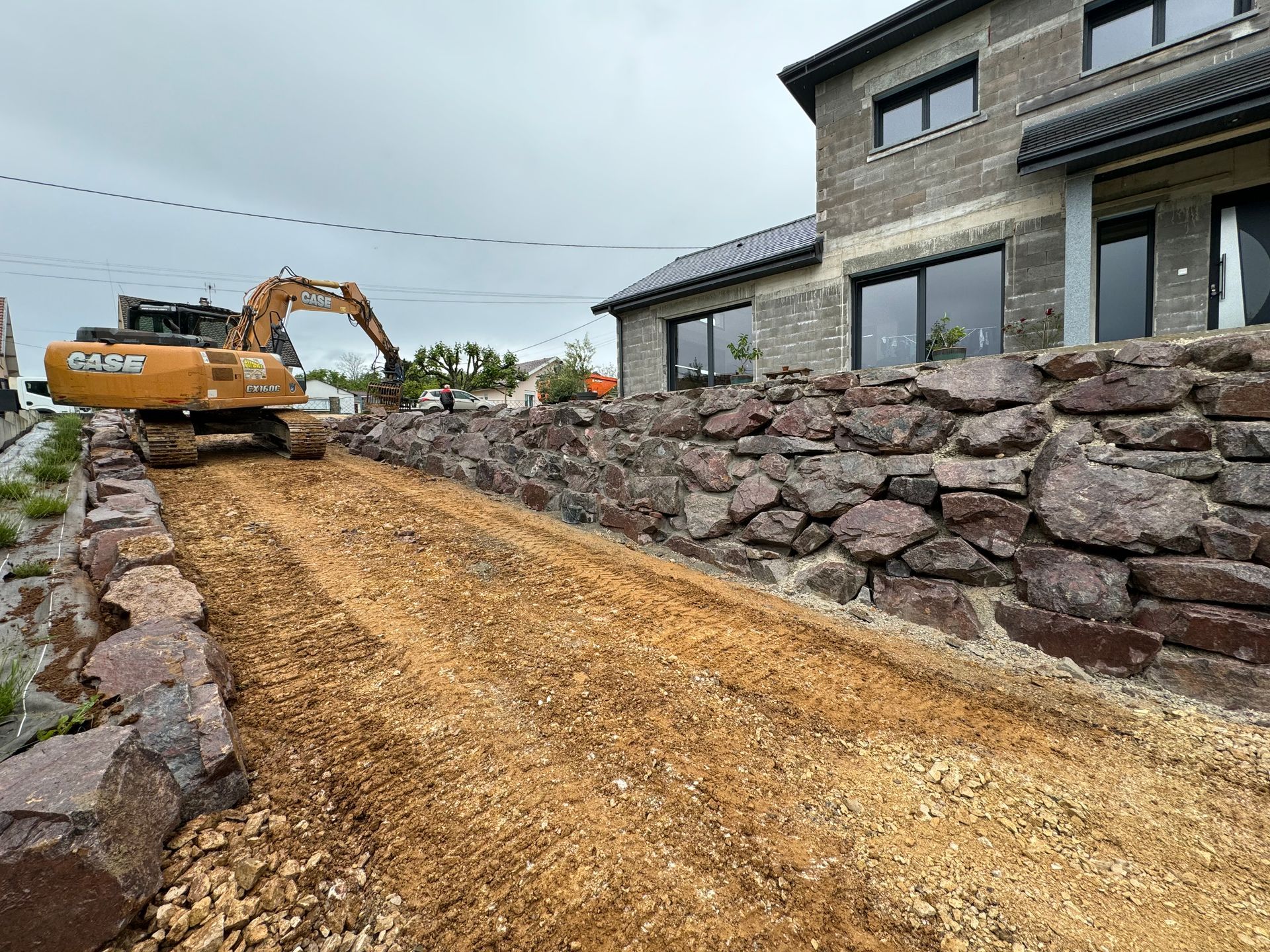 Une excavatrice sur une allée de gravier à côté d'un mur de soutènement en pierre et d'une maison